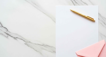 A gold pen lies on a blank white paper next to pink envelopes on a marble surfaceの写真素材