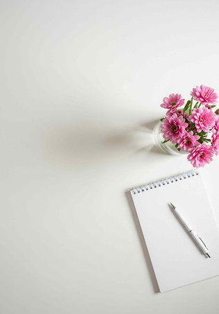 A minimalist workspace with a notepad, pen, and vase of pink flowers on a white desk