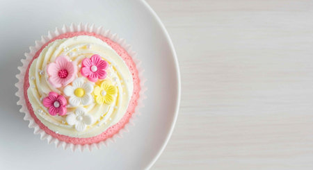 A delicious pink cupcake with white frosting and colorful flowers on top, placed on a white plate