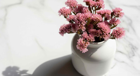 A white vase holds pink flowers on a marble surface with soft shadows.