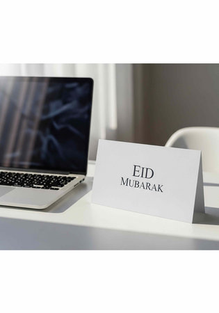 A white card with Eid Mubarak message on a desk next to a laptop