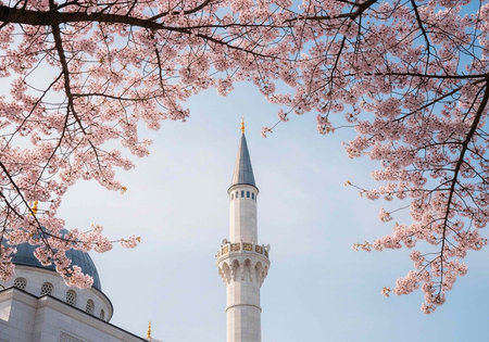 A white mosque tower stands tall under a clear blue sky surrounded by pink cherry blossoms.