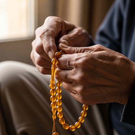 Elderly hands with wrinkles holding amber prayer beads near a window with natural light