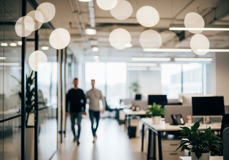 Blurred image of two men walking in a contemporary office space with desks, computers, and round...