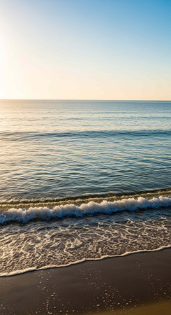 Calm ocean water with small waves washing over dark sand beach under a clear blue skyの写真素材