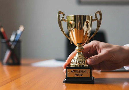 Hand holding shiny gold trophy on office desk with blurred background and achievement award plaque