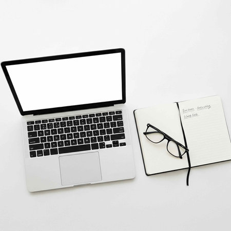 A silver laptop with black keyboard and glasses on a notebook against a plain white background