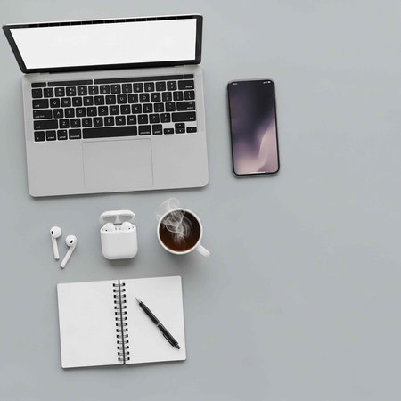Flat lay of office essentials including laptop, phone, earbuds, notebook, and coffee cup