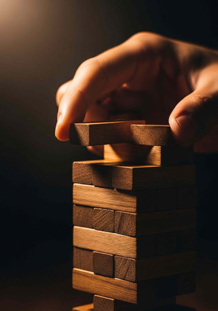 A hand carefully placing a wooden block on top of a precariously stacked tower in a dark room