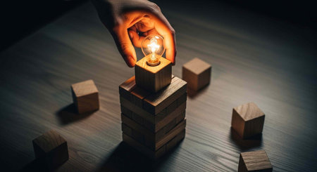 A hand holds a lit bulb on stacked wooden blocks with scattered cubes on a dark surface
