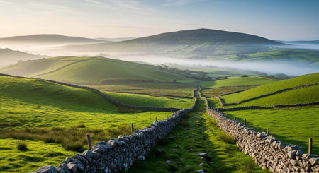 Lush green landscape with stone walls, rolling hills, and misty valleys under a clear blue sky