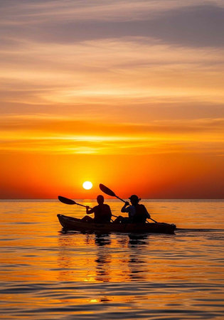 Two people kayaking on calm water during a vibrant sunset with orange and yellow hues in the skyの写真素材