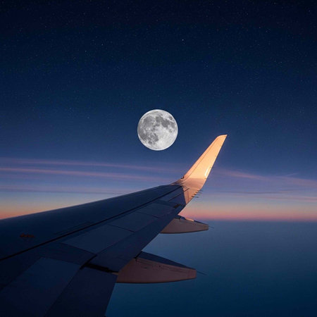 Airplane wing in flight under full moon with starry night sky and clouds at sunset
