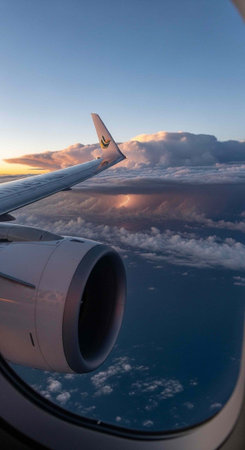 An airplane wing and engine are visible through a window as the plane flies above clouds at sunset
