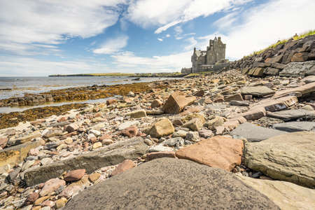 Historic hotel of Ackergill Tower at Sinclair Bay in the Scottish Highlands. Rocky beach with the beautiful and luxuary old british mansion in the background. Cloudy but sunny day at Wick, Scotland.のeditorial素材