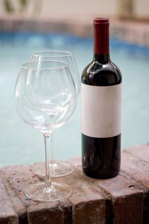 A red wine bottle and two empty glasses by the pool. Plenty of copy space on the blank wine label.の写真素材