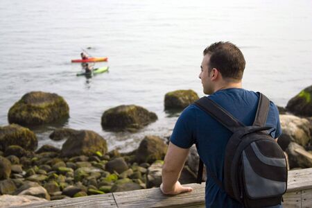 A young man enjoys the view by the sea as two people kayak in the distance.  Shallow depth of field.の写真素材