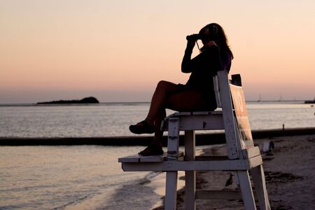 A silhouette of a woman looking with binoculars at the beach while seated in a lifeguard chair.の写真素材