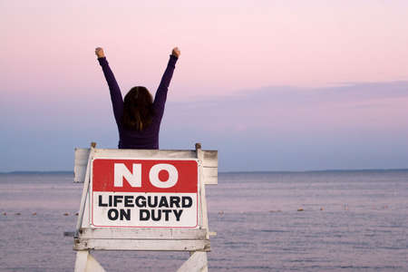 A woman holds her arms up in the air while seated in a vacant lifeguard chair at the beach.の写真素材