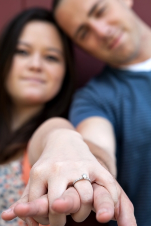 Young happy couple holding hands and showing the diamond engagement ring.  Shallow depth of field with strongest focus on the ring.の写真素材