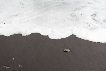 Close-up bisected image with white foam surf to top and black sand to bottom with several random pebblesの写真素材