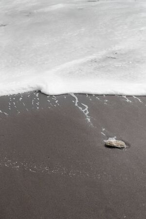 Close-up bisected image with white foam surf to top and black sand to bottom with single pebble to one sideの写真素材