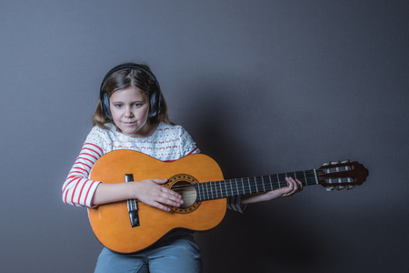 Teenage girl listening to music on headphone wearing white and red top and playing guitar sitting on a stool with a grey backgroundの写真素材