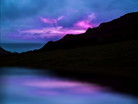 Dark hill on seashore and pink sunset reflecting in water