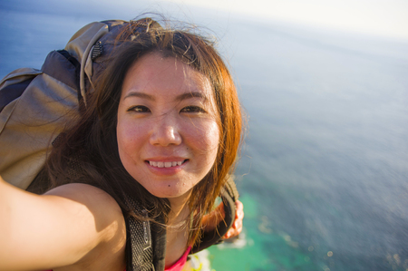 young beautiful and happy Asian Korean tourist woman taking selfie picture smiling carrying backpack after hiking excursion on top of tropical sea cliff landscape in holidays travel destinationの写真素材