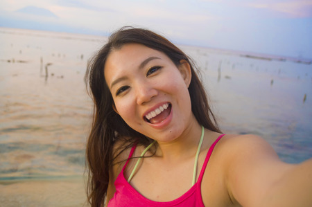 young happy and beautiful Asian Korean tourist woman taking self portrait selfie with mobile phone smiling having fun at sunset beach in digital nomad traveler blog and Summer holidays travelの写真素材
