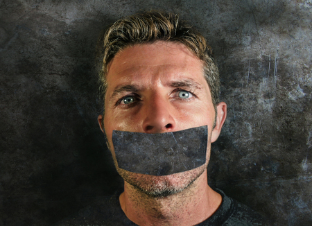 young man with mouth and lips sealed covered with adhesive tape in censorship coerced freedom of speech and forced silence and secrecy concept isolated on dark grunge backgroundの写真素材