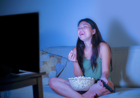 young pretty and happy Asian Korean woman sitting at living room sofa couch watching television...