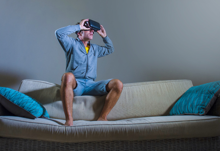 young attractive gamer man using VR goggles headgear technology playing simulator 3D video game having fun on home sofa couch enjoying exciting experience playing with virtual reality glasses の写真素材