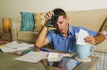 young attractive worried and stressed man at home calculating month tax expenses with calculator accounting payments doing bank paperwork in stress looking desperate suffering financial problem  の写真素材