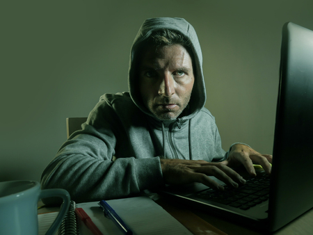 dangerous looking young hacker man in hoodie typing on laptop computer hacking and decoding system data or having illegal access breaking password on cyber attack and internet security conceptの写真素材