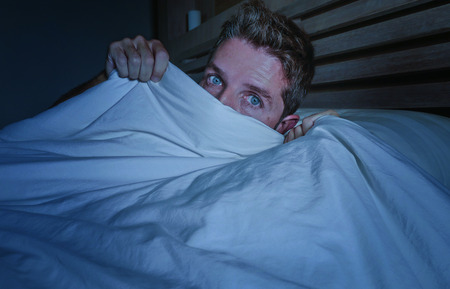 portrait of young attractive scared man in fear and panic suffering horror nightmare covering face with blanket sleepless at night lying on bed in dark bedroom in sleeping disorder conceptの写真素材