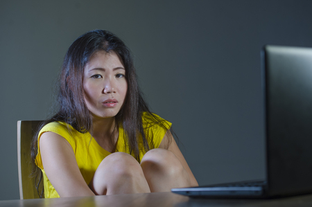young pretty scared and overwhelmed Asian Korean girl looking stressed at laptop computer feeling depressed and frightened in cyber bullying concept being stalked on internet social media の写真素材