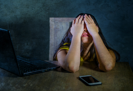 dramatic portrait of young latin sad and scared woman covering face with hands desperate and depressed with laptop and mobile phone in cyber bullying and internet problem の写真素材