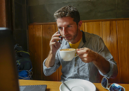 young attractive happy and successful millennial man working from internet coffee shop with laptop computer talking on mobile phone relaxed in entrepreneur modern digital nomad job の写真素材