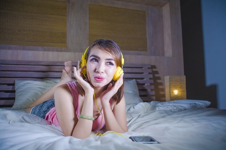 young attractive and happy woman in bed with yellow headphones listening to music on internet with mobile phone dancing and singing relaxed and cheerful in audio technology and lifestyle conceptの写真素材
