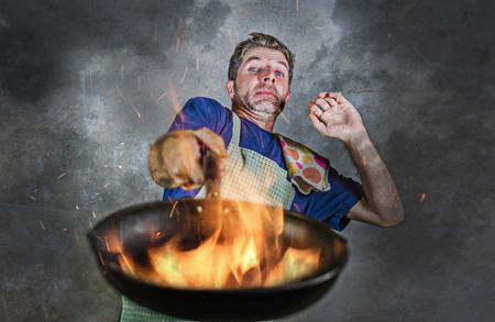 young attractive and shocked messy home cook man with apron holding pan in fire burning the food in kitchen disaster and unskilled and unexperienced terrible home cook at domestic cookingの写真素材