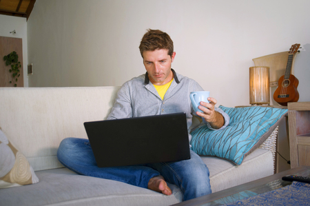 young attractive and successful man working from home living room sitting on couch networking on laptop computer drinking coffee happy in freelancer and independent job business conceptの写真素材