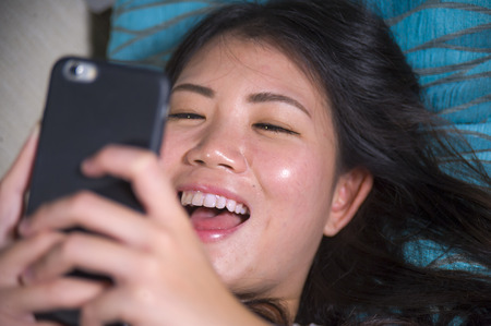 young sweet happy and pretty Asian Korean girl using internet social media app on mobile phone smiling cheerful lying on home sofa couch in online communication lifestyle conceptの写真素材