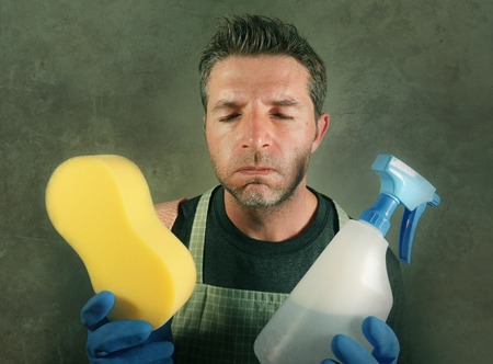 young tired and frustrated single man or house husband cleaning with detergent spray and sponge...