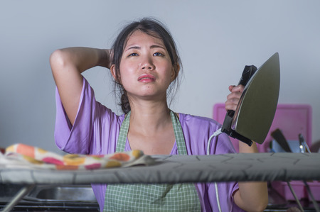 young pretty frustrated and stressed Asian Chinese woman with iron working at home kitchen ironing clothes desperate and overwhelmed in house maid service housework domestic stressの写真素材