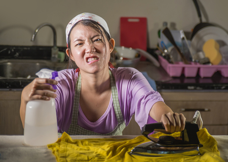 young pretty frustrated and stressed Asian Chinese woman with iron working at home kitchen ironing clothes desperate and overwhelmed in house maid service housework domestic stressの写真素材