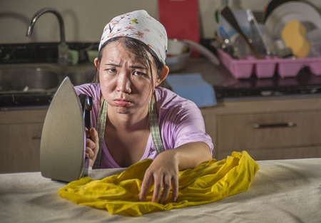 portrait of young pretty frustrated and stressed Asian Korean woman working at home kitchen ironing clothes desperate and overwhelmed in house maid service housework and domestic stressの写真素材
