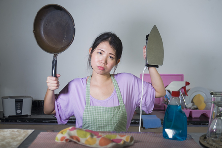 young beautiful and frustrated Asian Korean woman holding pan and iron stressed and upset overwhelmed by domestic work and chores at home kitchen in housekeeping stress conceptの写真素材