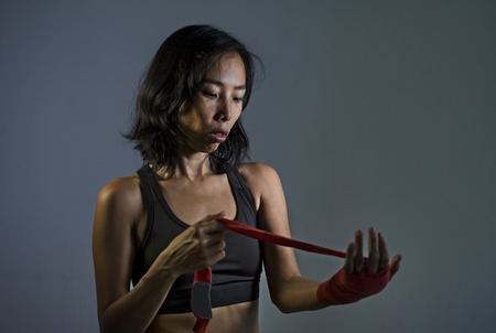 portrait of sporty and fit fighter Asian Chinese woman using wrist wraps wrapping before mma fight sport or boxing workout looking dangerous in badass pose isolated on dark grungeの写真素材