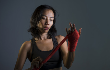 portrait of sporty and fit fighter Asian Chinese woman using wrist wraps wrapping before mma fight sport or boxing workout looking dangerous in badass pose isolated on dark grungeの写真素材